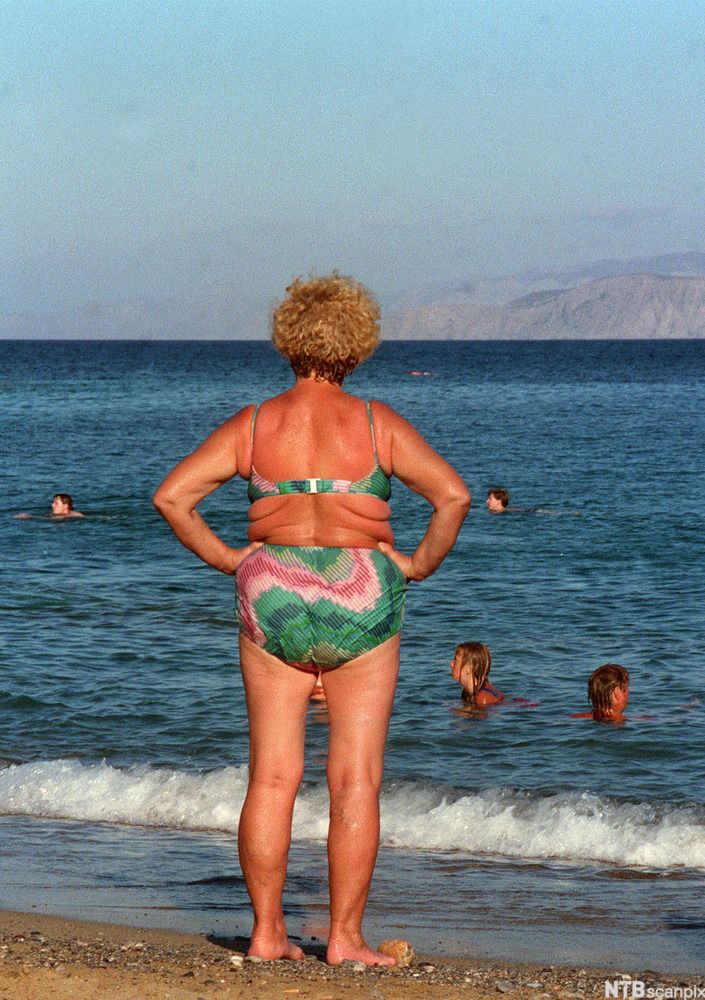 En middelaldrende, solbrun kvinne i bikini står med ryggen til og ser ut over havet. Foto. 