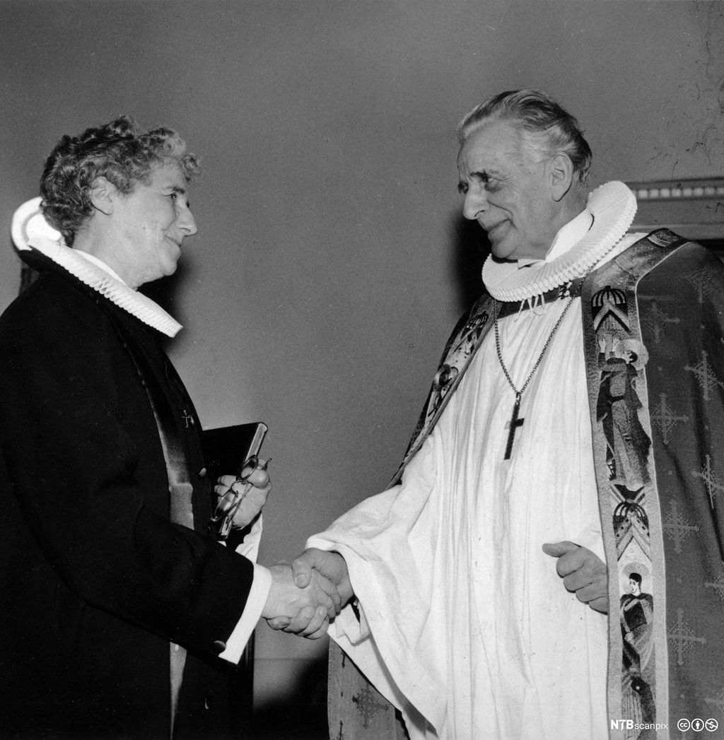 En kvinne i prestekjole og en mann iført biskopens embetsdrakt. De smiler til hverandre og tar hverandre i hånden. Svart/hvitt-foto.