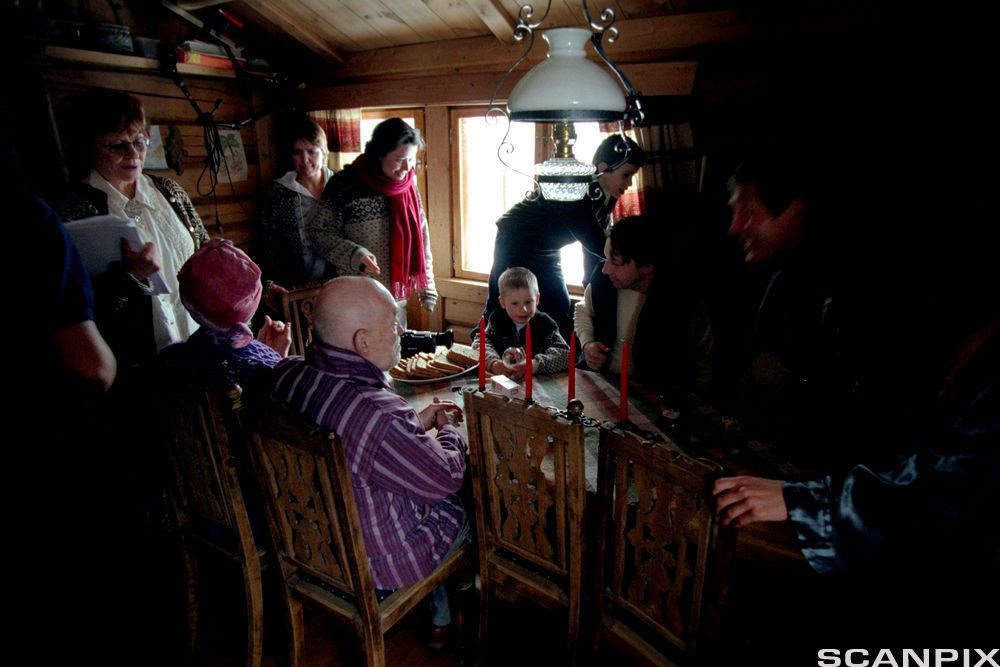 Flere personer samlet i et lite, trangt rom med tømmervegger. Noen sitter ved et spisebord, mens andre står eller beveger seg ved bordet. Foto.