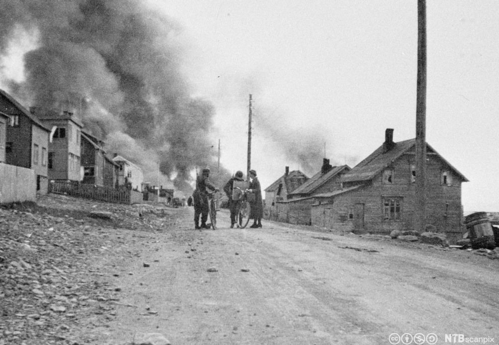 Soldater midt i gata i et boligområde i en liten by. Det brenner og ryker i bakgrunnen. Svart-hvitt-foto. 