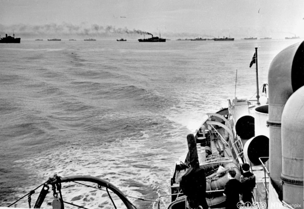 Flere større og mindre skip på rekke i konvoi ute på havet. I forkant er et skip med norsk flagg og noe mannskap på dekk. Svart-hvitt foto. 