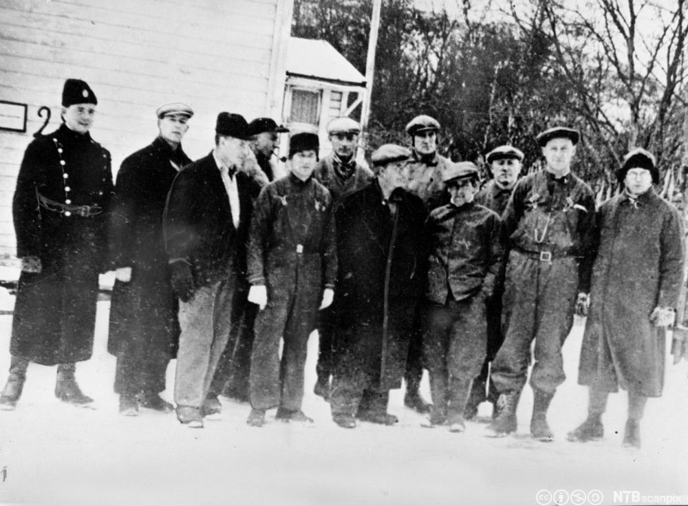 Elleve menn i forskjellige klær står oppstilt i snøen foran et hus. Ved siden av står det en vaktmann i uniform. Foto.