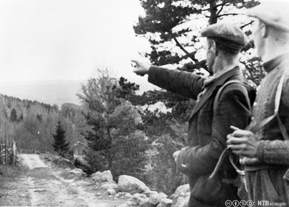 To menn med ryggsekk langs en skogsvei. Den ene mannen peker mot åsene i bakgrunnen. Svart-hvitt foto.