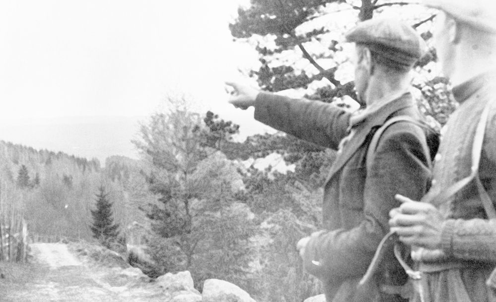 To menn med ryggsekk langs en skogsvei. Den ene mannen peker mot åsene i bakgrunnen. Svart-hvitt foto.