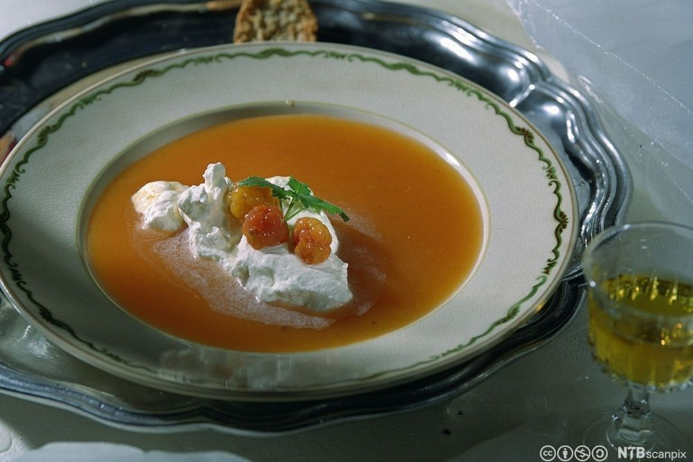 Oransje suppe med ein stor klatt rømme i midten og tre molter og eit grønt blad som garnityr på toppen. Foto.