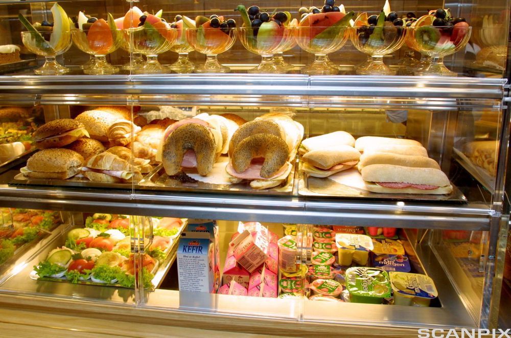 Glasdisk med kantinemat: fruktbeger, rundstykke, croissantar, horn med ost og skinke, panini med salami og ost, smørbrød, kefir, mjølk og jogurt. Foto.
