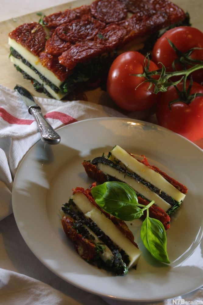 Lag av tomater og mozzarella presset sammen i avlange serveringsstykker. Anrettet på en hvit tallerken. Foto.
