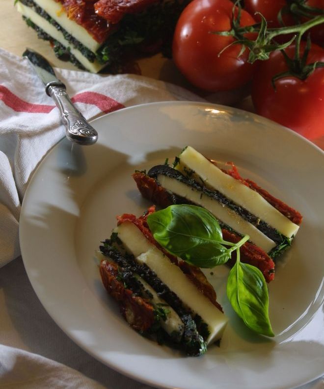 Lag av tomater og mozzarella presset sammen i avlange serveringsstykker. Anrettet på en hvit tallerken. Foto.