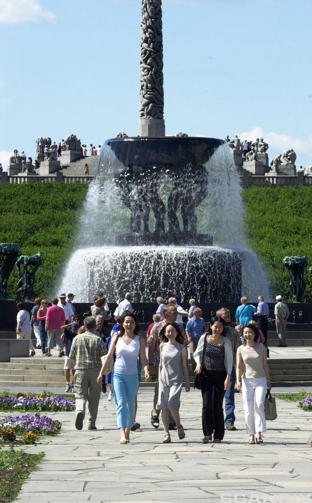 Fire smilende personer går i retning kamera. I bakgrunnen er det steinskulpturer og en vannfontene. Flere personer går blant skulpturene. Foto.