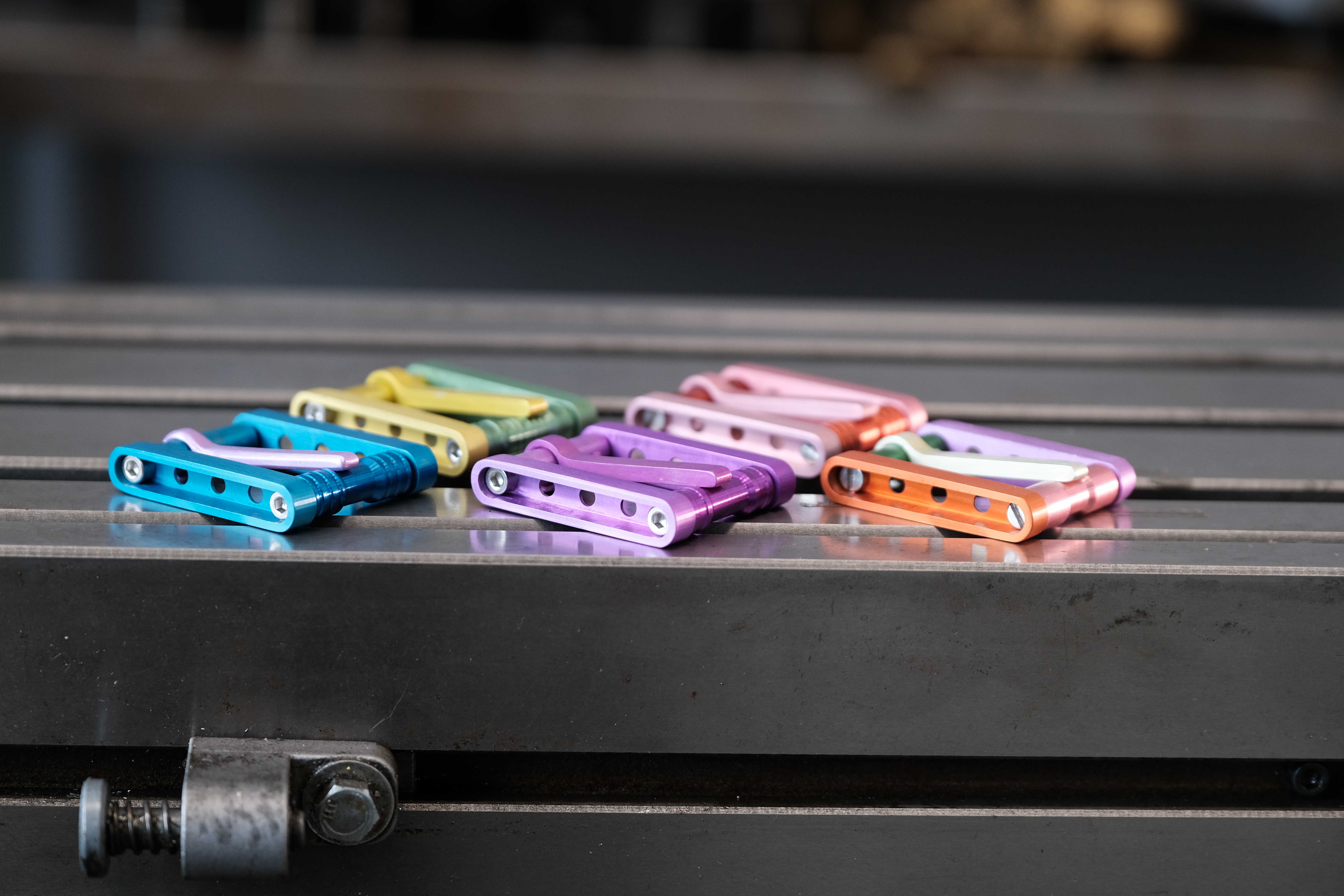 Fem beltespenner i ulike farger liggende på en arbeidsbenk. De er dels ensfarga, dels tofarga. Fargene er blått, grønt, gult, rosa, lilla og oransje. Foto.