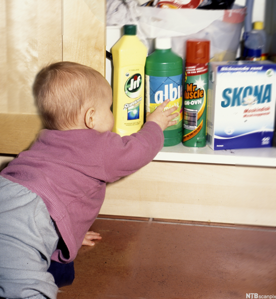 Baby griper etter en flaske vaskemiddel som står i et kjøkkenskap. Foto.