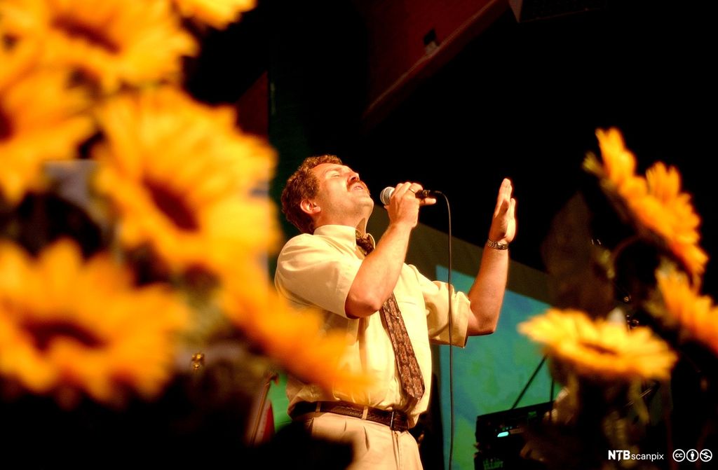 En engasjert mann med mikrofon taler til en forsamling. Foto.