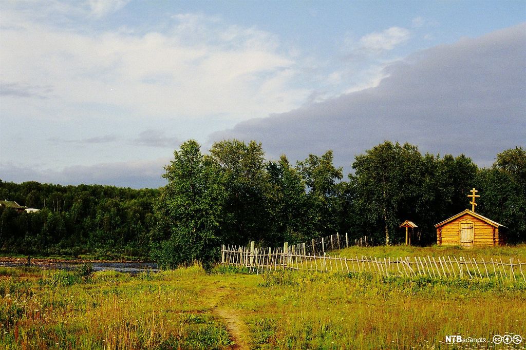 Liten trebygning med ortodoks kors på toppen på en eng ved en elv. Foto.