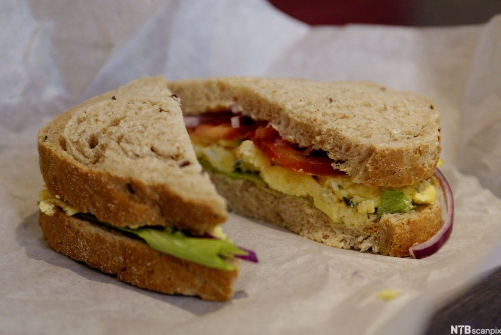 Dobbel brødskive delt i to, med løk, salat, tomat og egg, på et matpapir. Foto.