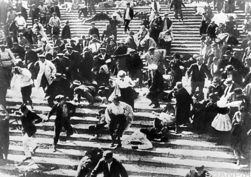 En menneskemengde på vei nedover ei lang og brei trapp. Noen løper, noen hopper, noen har falt, noen står stille. Svart-hvitt-fotografi.