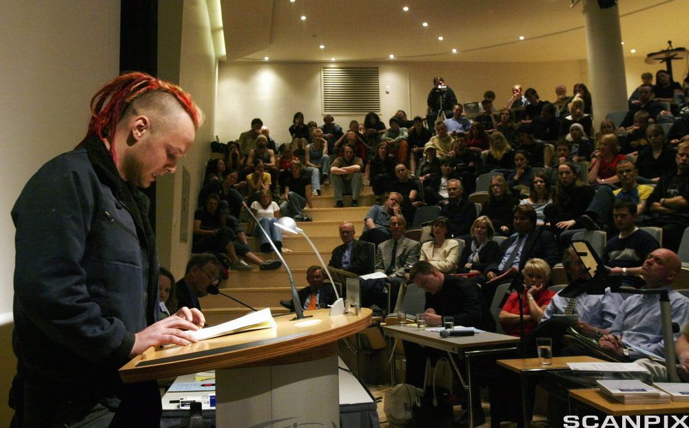 Ung mann holder innlegg i et auditorium med mange tilhørere. Foto.