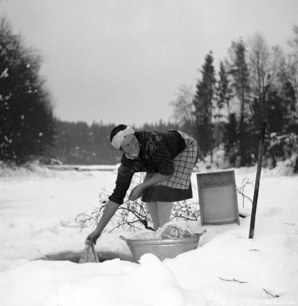Smilende kvinne i kjole og med oppbrettede ermer holder et plagg i vannet i et hull i isen. En metallbalje med klær og et vaskebrett står ved siden av henne. Svart-hvitt foto.