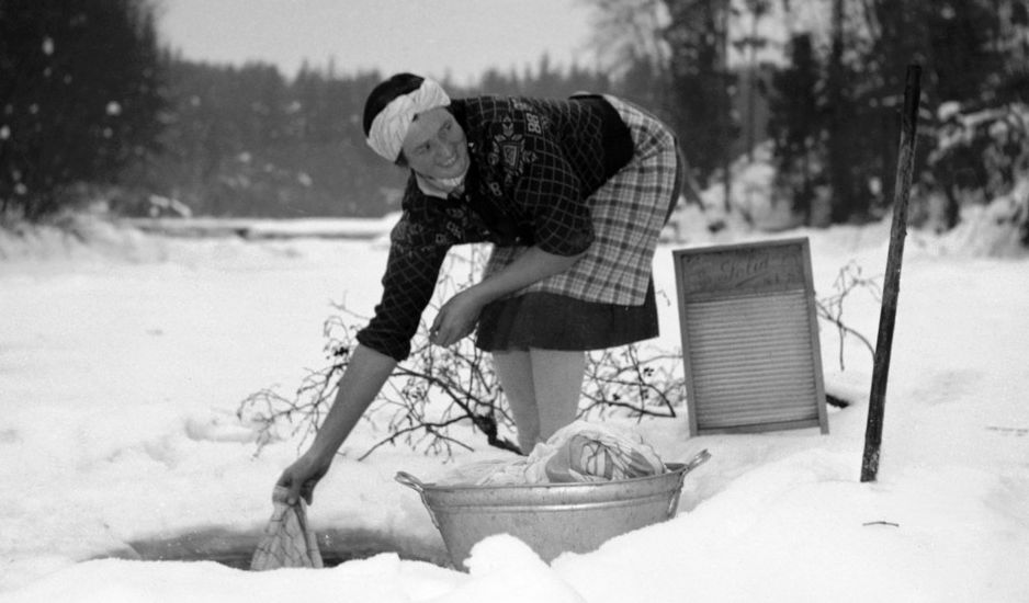 Kvinne vasker klær i et hull i isen i en bekk. Foto.