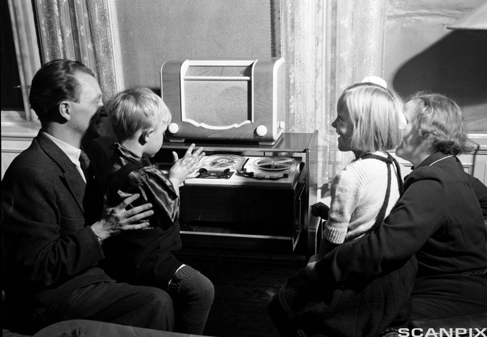 Familie med mor, far og to barn sitter foran radioapparatet i typisk 1950-tallsstue. Foto.