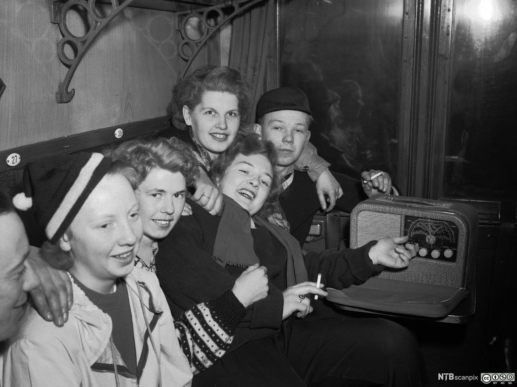 En gjeng glade ungdommer sitter i en togkupé og lytter til en reiseradio av merket Kurer. To av ungdommene røyker sigaretter. Foto.
