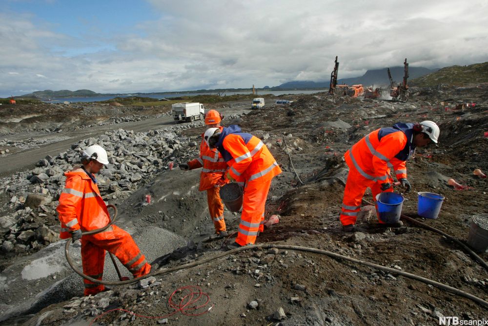 Fire arbeidere og ulike maskiner på en anleggsplass. Det foregår sprenging og flytting av løsmasse. Foto.