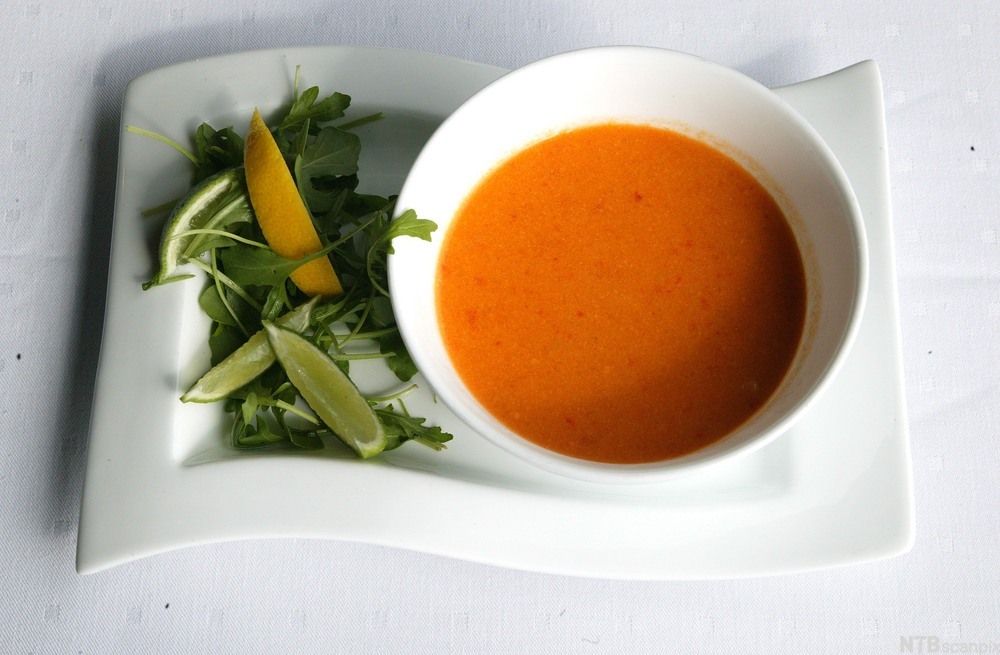 Oransje suppe i en bolle og grønnsaker anrettet på et hvitt fat ved siden av. Foto.