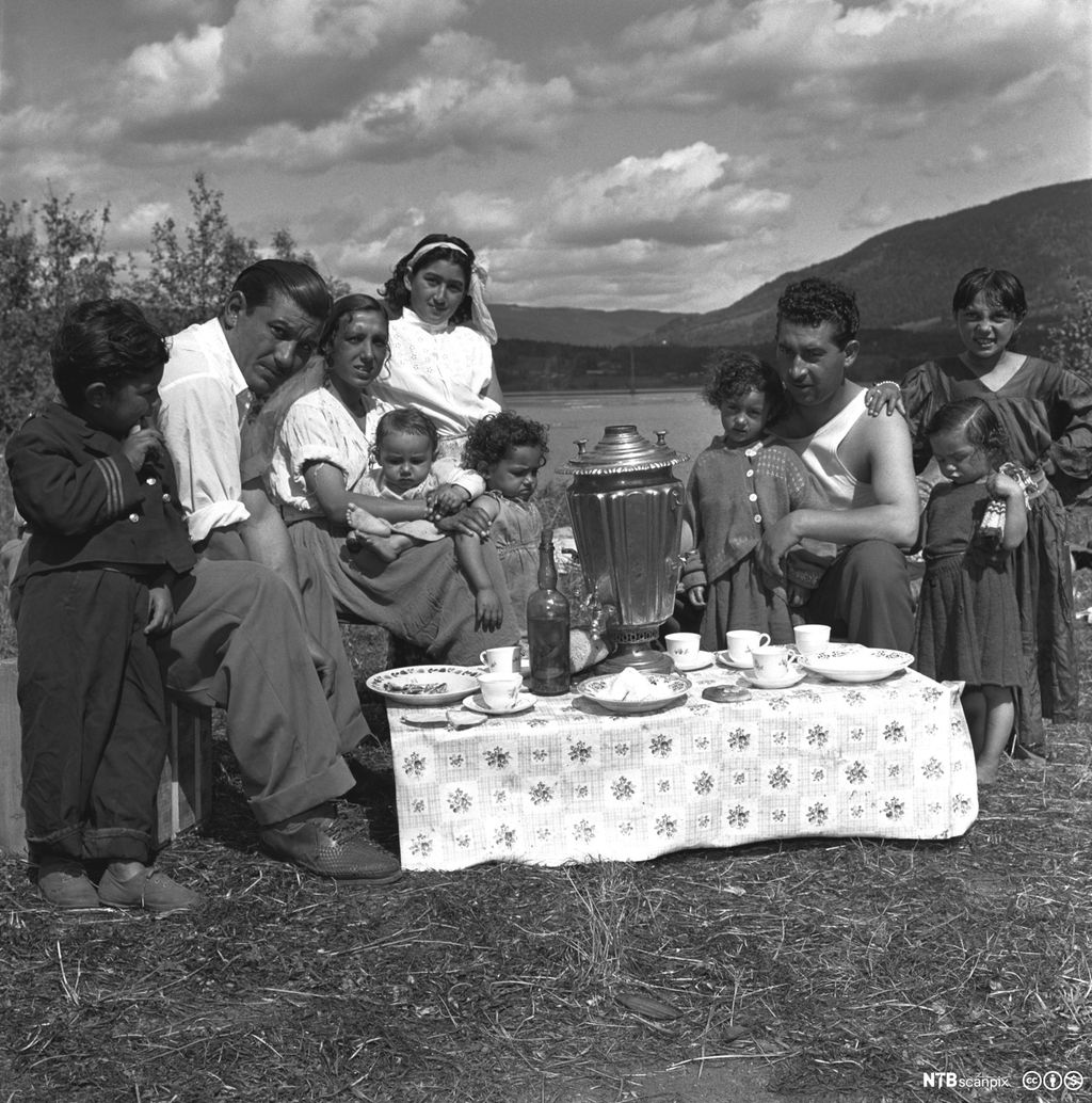 En stor familie spiser i det fri. Det er dekket på med duk på et lite bord. Svart-hvitt foto. 