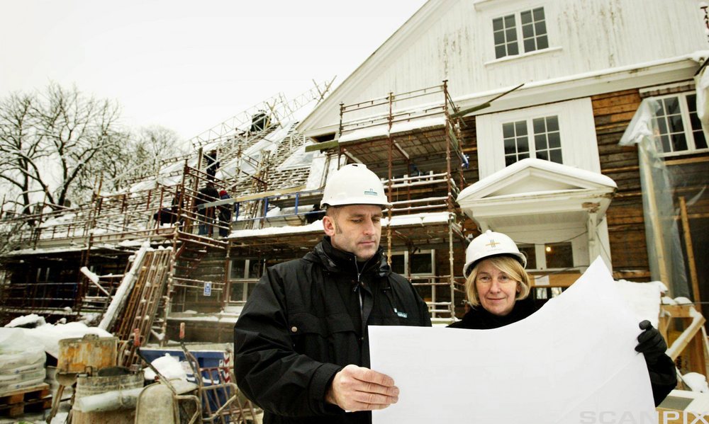 En mann og en kvinne med hjelm og arbeidstøy står på en byggeplass og studerer ei byggtegning. Foto.