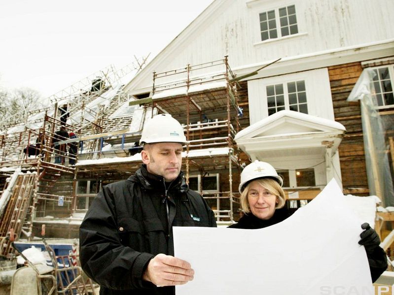En mann og en kvinne med hjelm og arbeidstøy står på en byggeplass og studerer ei byggtegning. Foto.