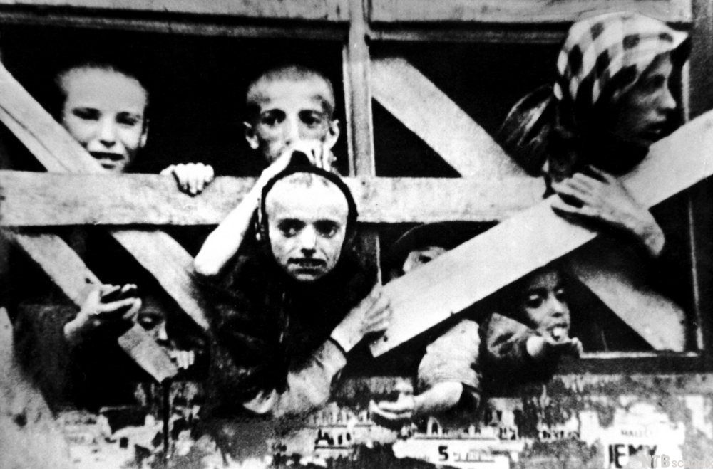 Barn i konsentrasjonsleir, sittende og liggende i ei sovebrakke. Sort-hvitt foto.