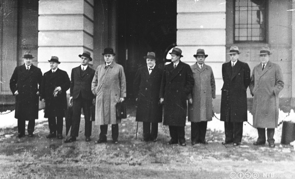 En rekke menn med frakk og hatt på slottsplassen. Svart-hvitt foto.