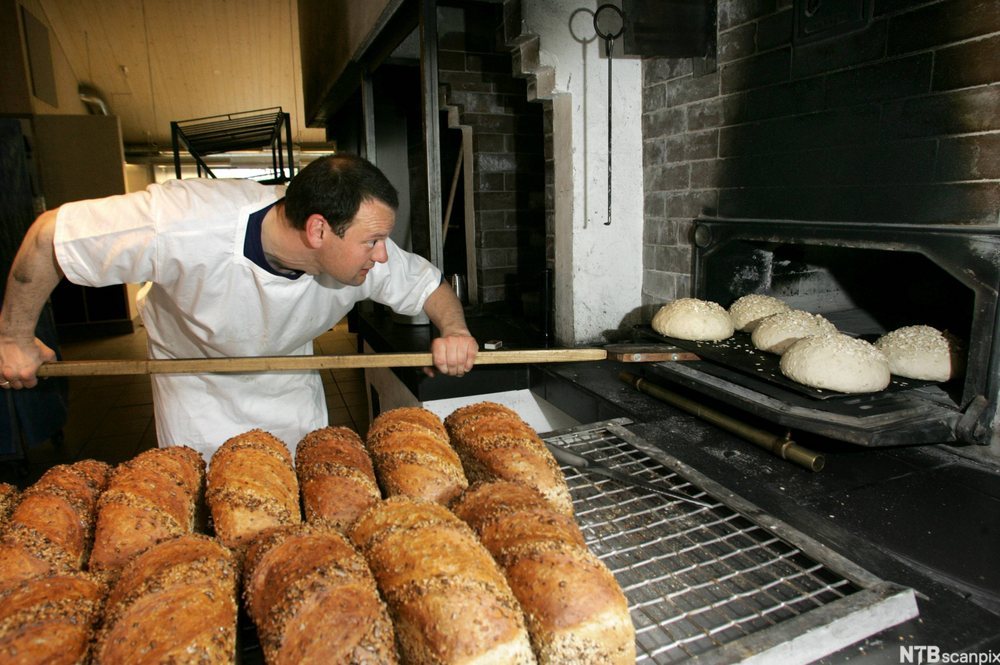 Mann plasserer runde brød i en bakerovn med en stor brødspade. Ferdige brød i forgrunnen. Foto.