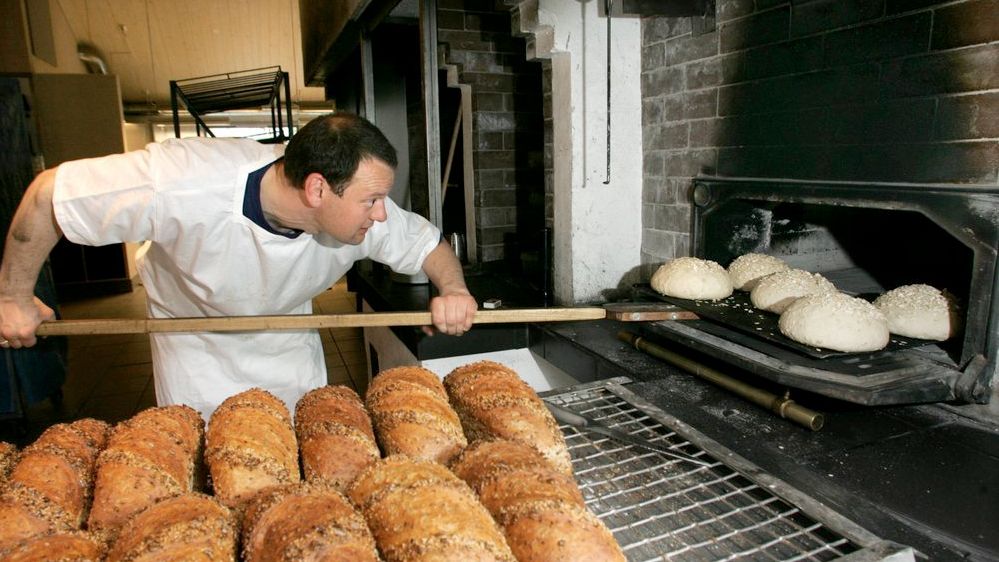 Mann plasserer runde brød i en bakerovn med en stor brødspade. Ferdige brød i forgrunnen. Foto.