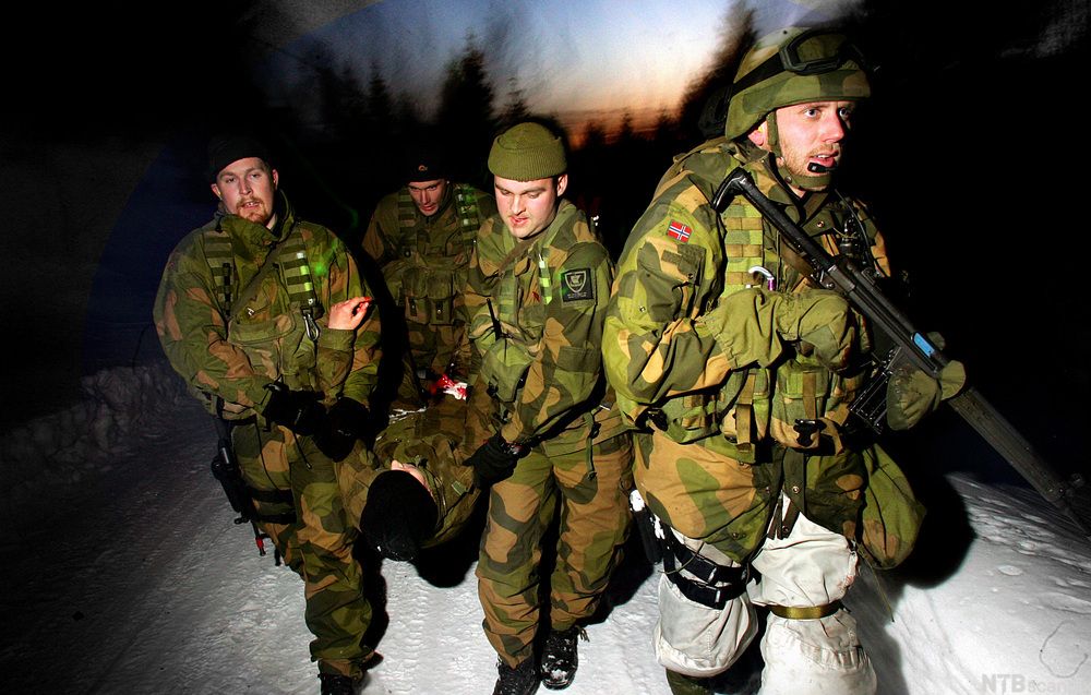 Tre menn i kamuflasjedrakter bærer en person mellom seg. En fjerde soldat går foran til høyre i bildet med et gevær i hendene. Det er natt og snø. Foto.