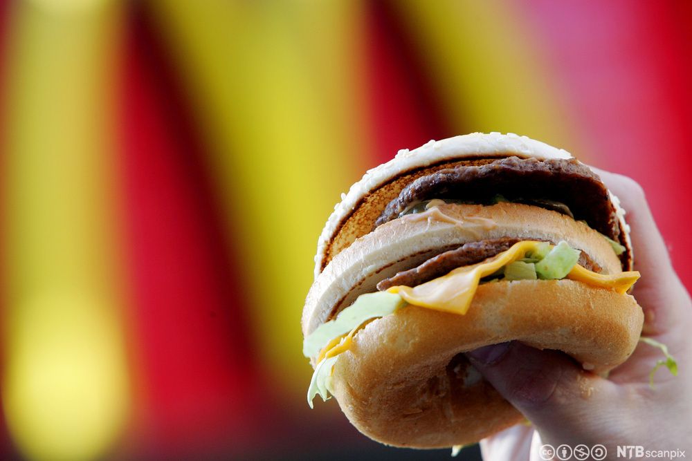 En hånd holder en dobbel cheeseburger med salat og dressing. I bakgrunnen kan man skimte den gule og røde logoen til McDonald's. Foto.