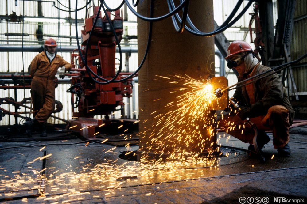 Sveisearbeid på oljeplattform i Nordsjøen, 1982. En arbeider i kjeledress, hjelm og sveisebriller sveiser så gnistene spruter. I bakgrunnen ser vi maskineri, kabler og en annen arbeider. Foto.