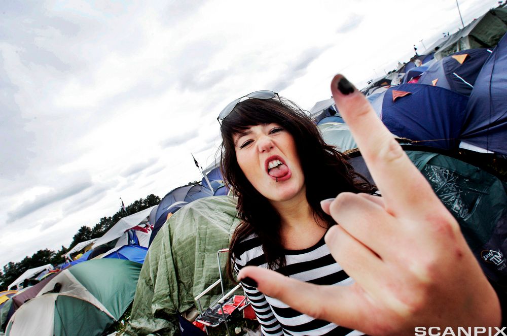 Ung kvinne står ute blant mange telt, ho rekker tunge til kameraet og viser djevelhornteikn med den eine handa. Foto.