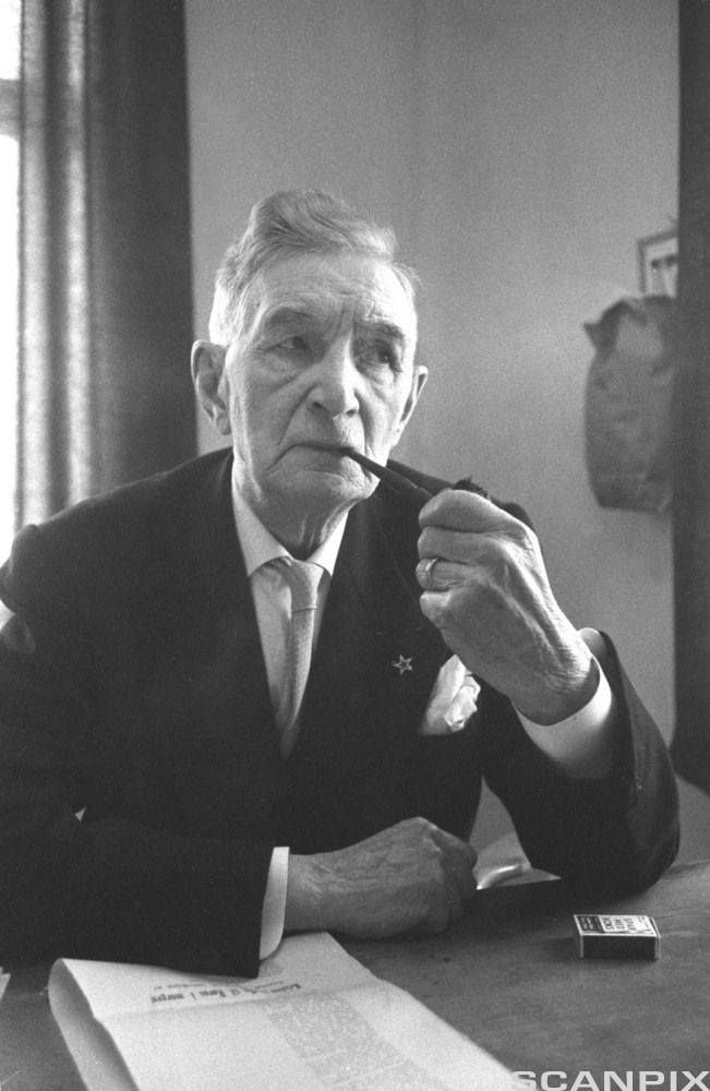 Eldre mann i dress sit ved eit bord og røyker pipe. Svart-kvitt foto.