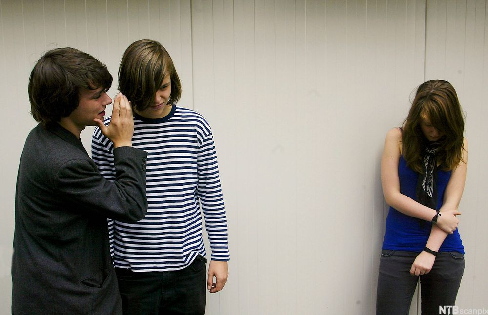 Ei jente står for seg selv og er trist. To gutter står litt lenger borte og hvisker til hverandre. Foto.