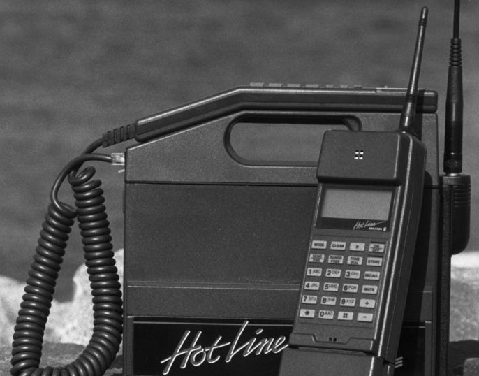 En gammel mobiltelefon formet som en boks med bærehåndtak. Det er en antenne på den, og det står HotLine skrevet på den. Svart/hvitt-foto.