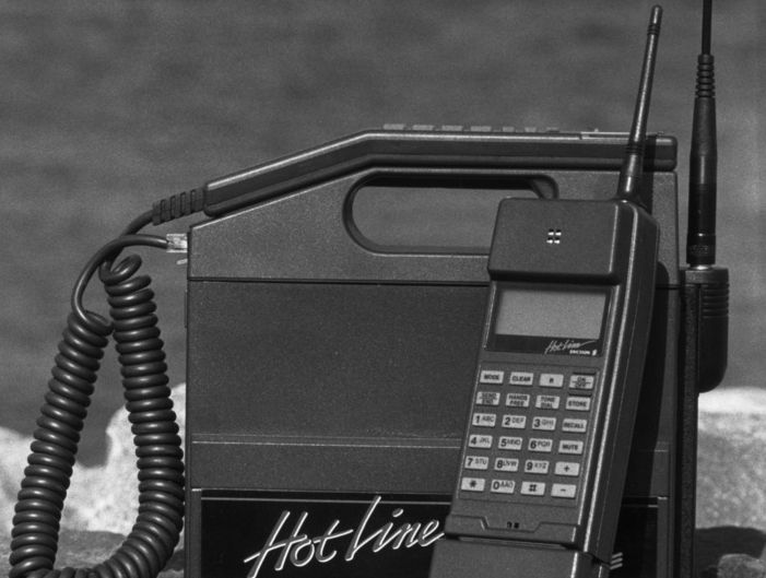 Ein gammal mobiltelefon forma som ein boks med berehandtak. Det er ei antenne på han, og det står HotLine skrive på han. Svart/kvitt-foto.