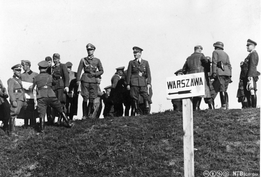 Flere menn i uniformer står på toppen av en grasbakke. Det er et skilt i forkant hvor det står Warszawa og en pil som viser retningen dit. Mannen i midten er Adolf Hitler. Foto. 