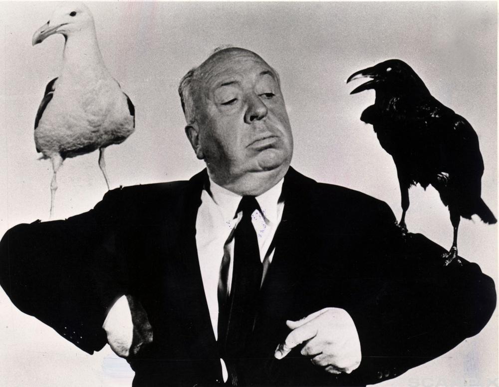 Alfred Hitchcock er kledd i svart dress og står med en hvit måke og en svart ravn på skuldrene. Foto.