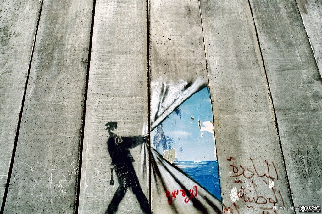 På den grå muren som skiller palestinere og israelere på Vestbredden er det malt et bilde av en politimann som lager hull i muren. Gjennom hullet ser en blått hav og himmel. Foto.