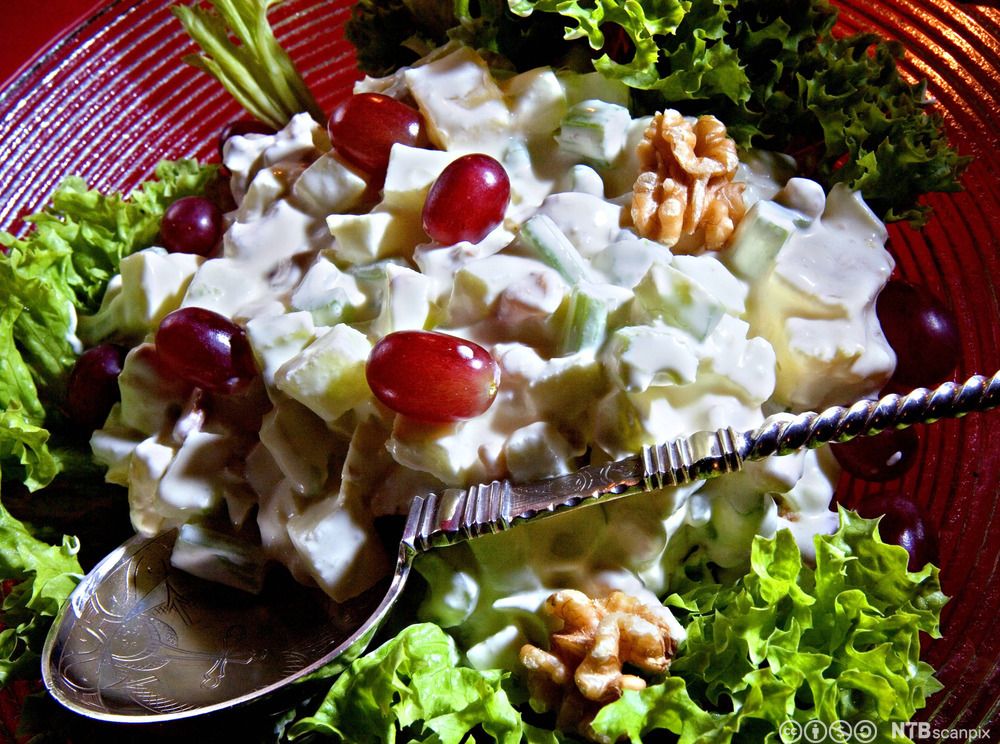 Rød serveringsbolle med waldorfsalat. I bunnen ligger grønne salatblader, og salaten er dekorert med røde druer og brune valnøtter. Foto.