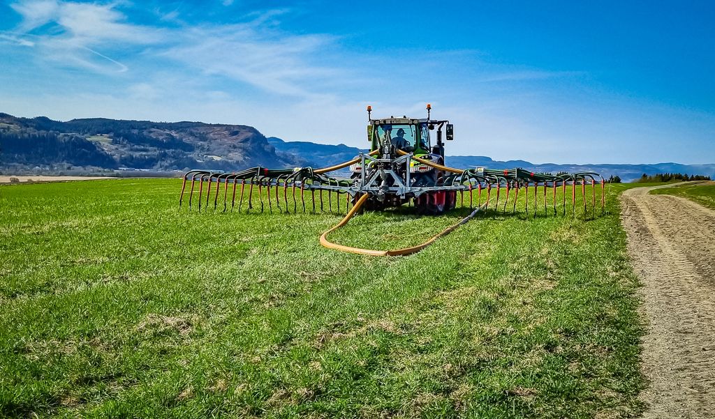 I trepunkten til en traktor henger redskap med en bred bom på tvers med slanger og rør. Traktoren kjører på et grasfylt jorde, og bak på utstyret er det koplet til en tjukk slange som ligger på bakken. Slangen strekker seg nedover jordet. Langs bommen henger det slanger ned mot graset. Foto.