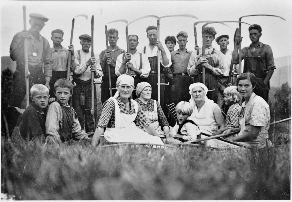 Kvinner og barn sitter i gresset. Bak dem står en rekke med menn med ljå. Svart-hvitt-foto.