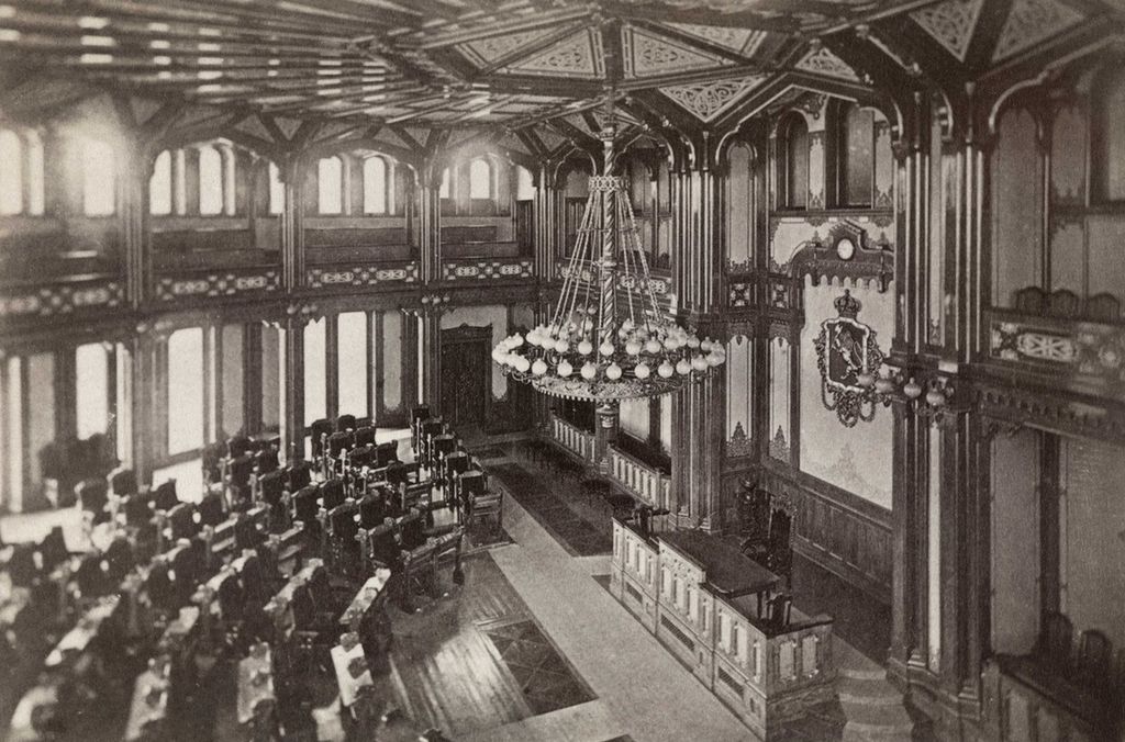 Stortingssalen med representantenes seter i halvsirkel og med stortingspresidentens stol og talerstolen fremst. Bildet viser salen slik  den så ut rundt 1870. Foto.