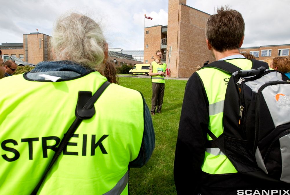 Ei gruppe mennesker står ute og hører på en mann som snakker. De har gule vester med påskriften "streik". Foto.