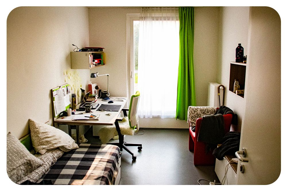 Studenthybel. Rommet har ei dør, et skap, en liten kommode, en lenestol, to vegghyller, et stort vindu med lange gardiner, et skrivebord, en svingstol og ei seng. På skrivebordet står det ei lampe. Foto.